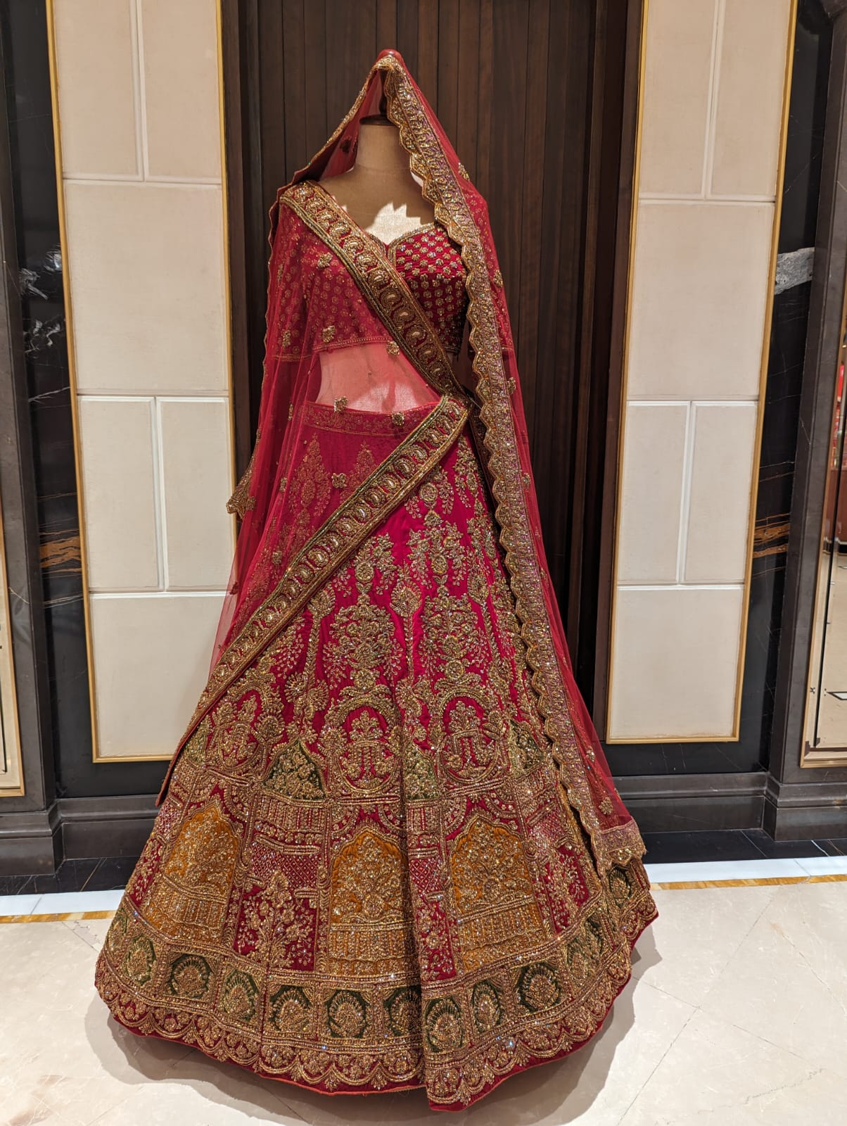 Traditional embroidered lehenga with red and gold patterns on a mannequin against a tiled wall.