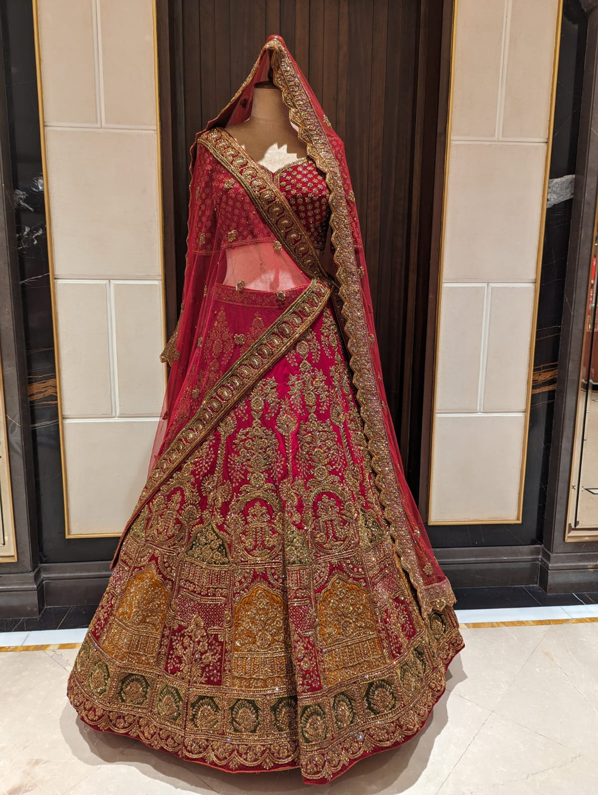Traditional embroidered lehenga with red and gold patterns on a mannequin against a tiled wall.