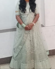 A woman wearing a light sage green embroidered traditional outfit with a matching dupatta, standing indoors against a plain white wall. Her face is obscured by a yellow smiling emoji, and a brass oil lamp is visible on the right side.