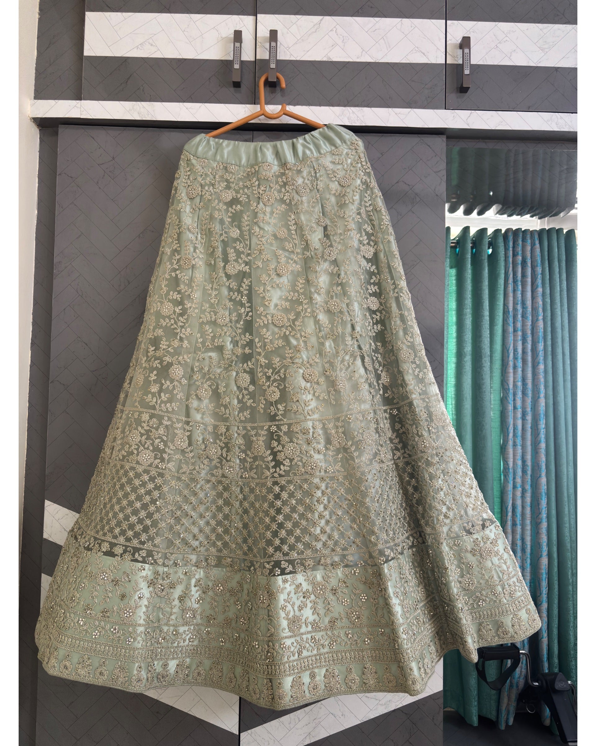 An olive green embroidered lehenga skirt with intricate metallic gold and silver floral patterns, hanging on a wooden hanger against a gray and white paneled wardrobe door.