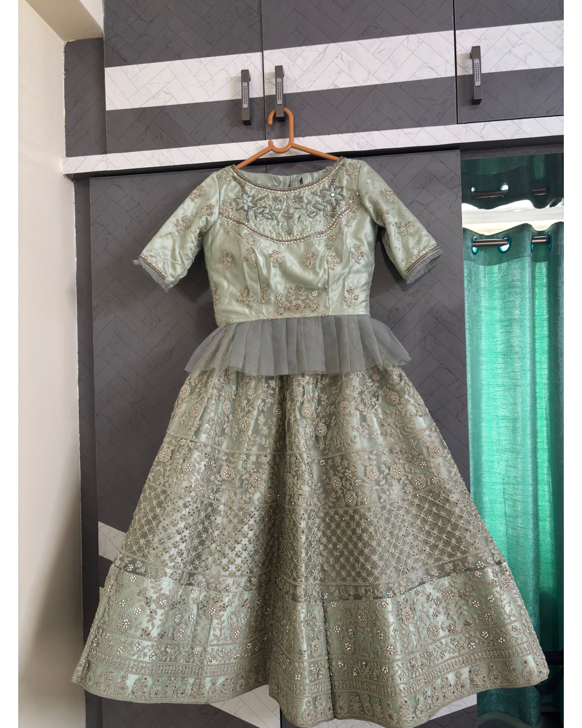 A light sage green embroidered traditional Indian dress with a sheer tulle waistband, hanging on a wooden hanger against a dark gray wardrobe door.
