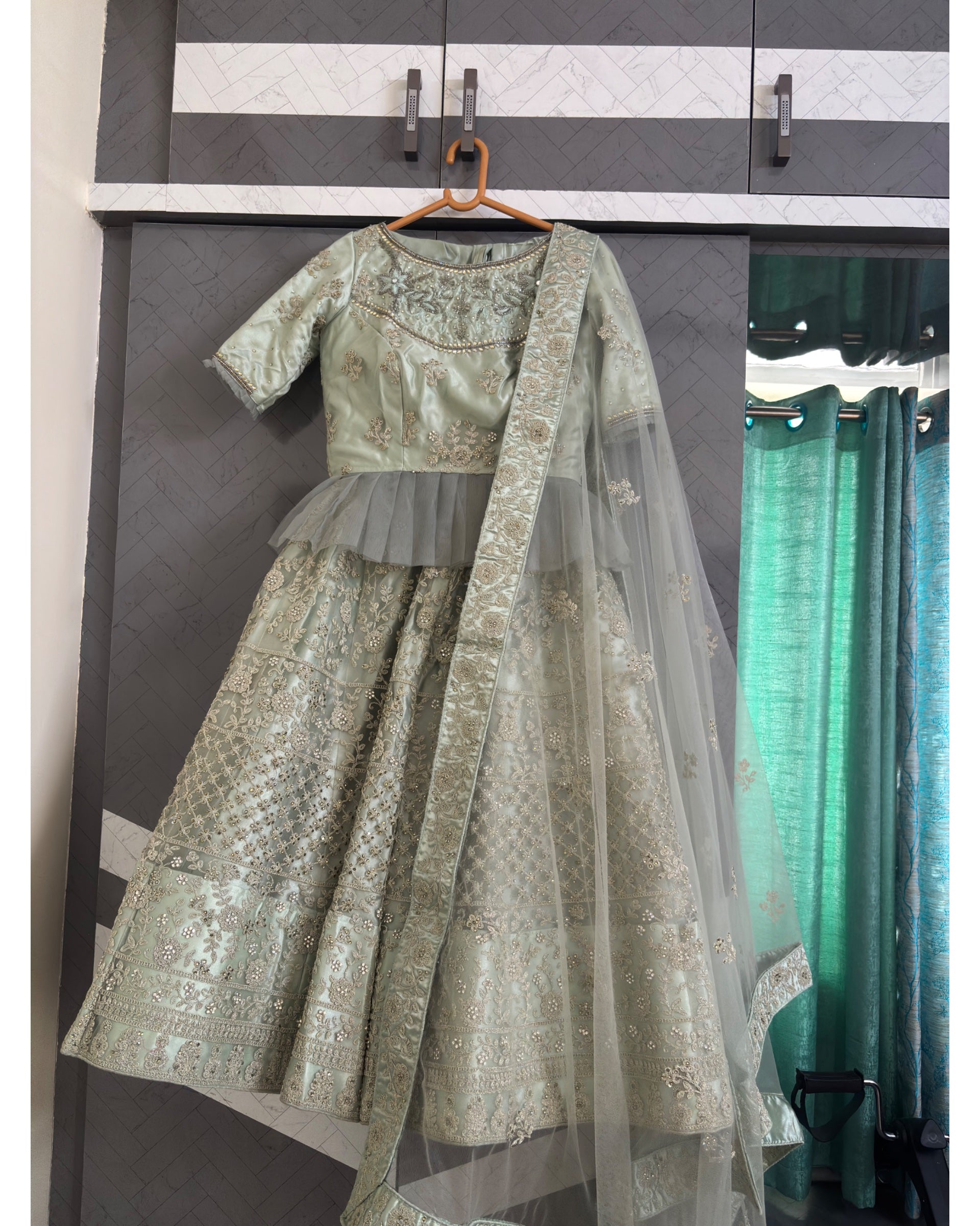 A light sage green embroidered traditional Indian lehenga choli set with a matching dupatta, hanging on a wooden hanger in front of a gray wardrobe door.