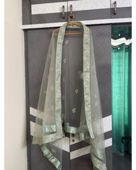 A light sage green sheer dupatta with gold floral embroidery and a matching metallic border, draped over a dark gray dress form on a wooden hanger in front of a patterned gray wardrobe.