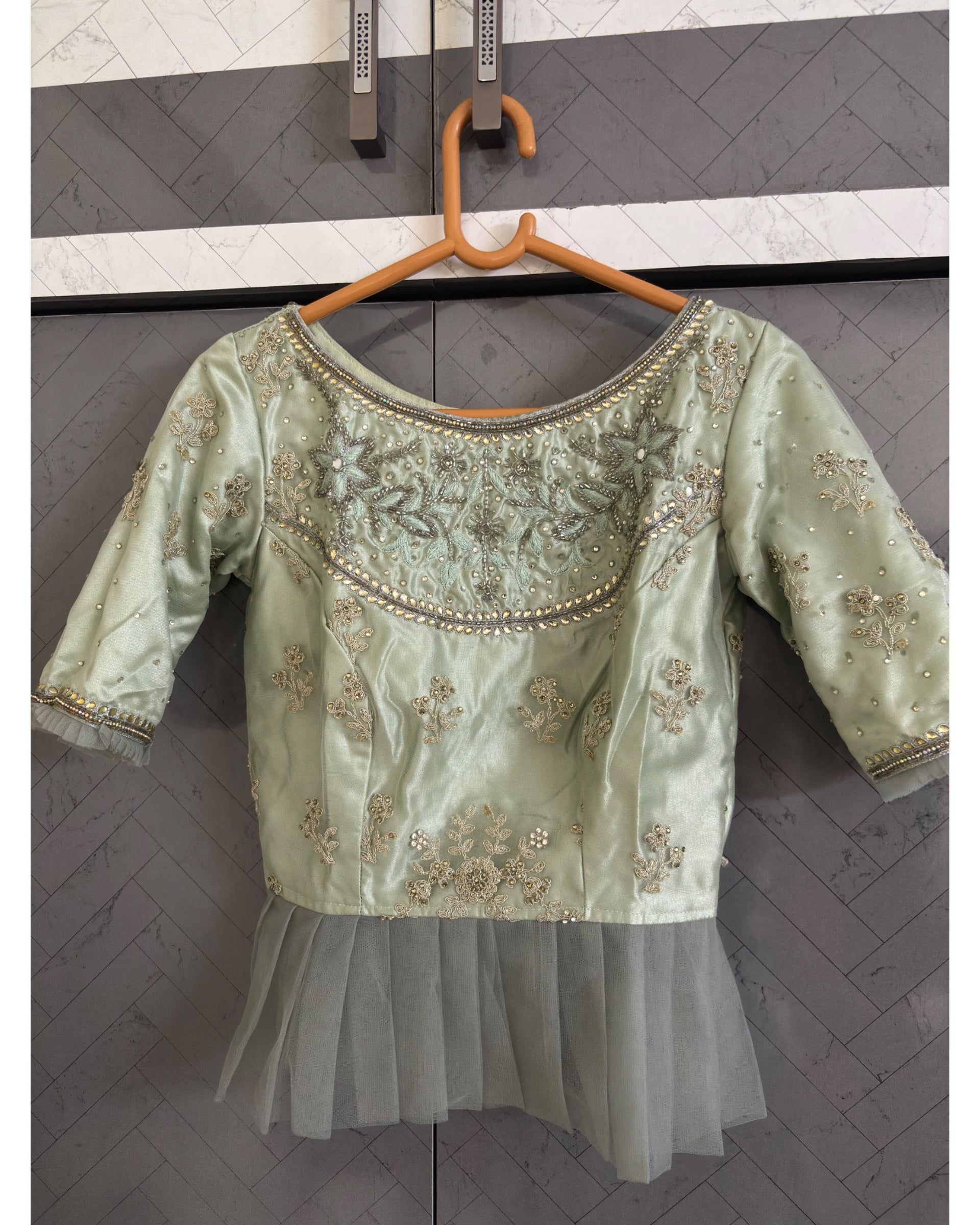 A light sage green blouse with intricate gold and silver embroidery, displayed on a wooden hanger against a gray paneled wall.