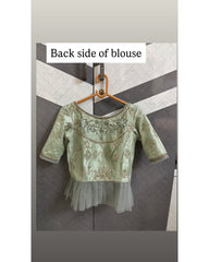 A light sage green embroidered blouse, shown from the back on a wooden hanger against a gray paneled door. The text overlay at the top reads 'Back side of blouse'.