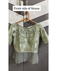 A light sage green embroidered blouse on a wooden hanger, featuring a satin-like bodice with gold and silver mirror work and bead borders, a sheer mesh overlay at the hem, and three-quarter sleeves. A text label at the top reads 'Front side of blouse'.