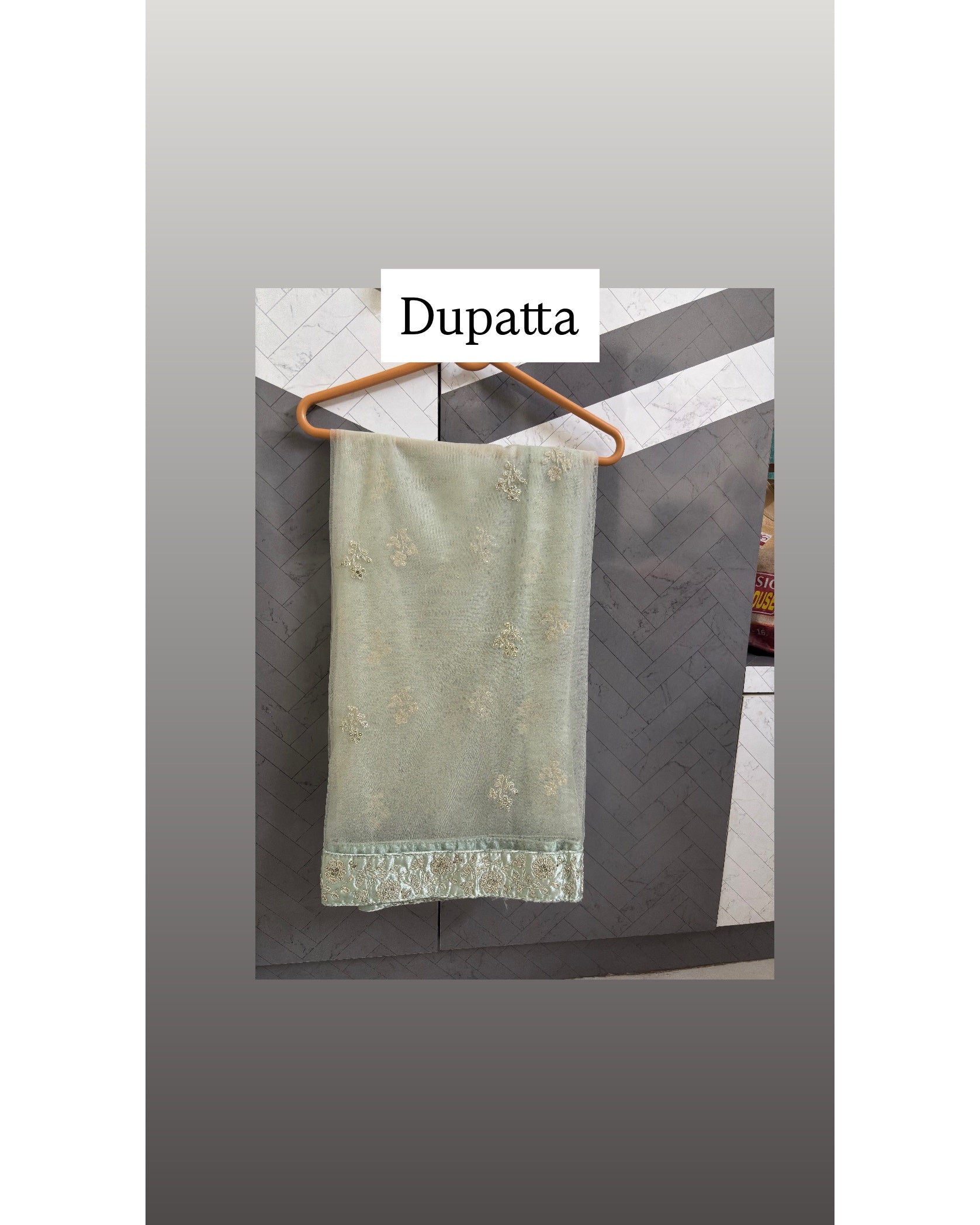 A sage green dupatta with delicate gold floral embroidery and a matching embroidered border, hanging on a wooden hanger against a dark gray paneled wall.