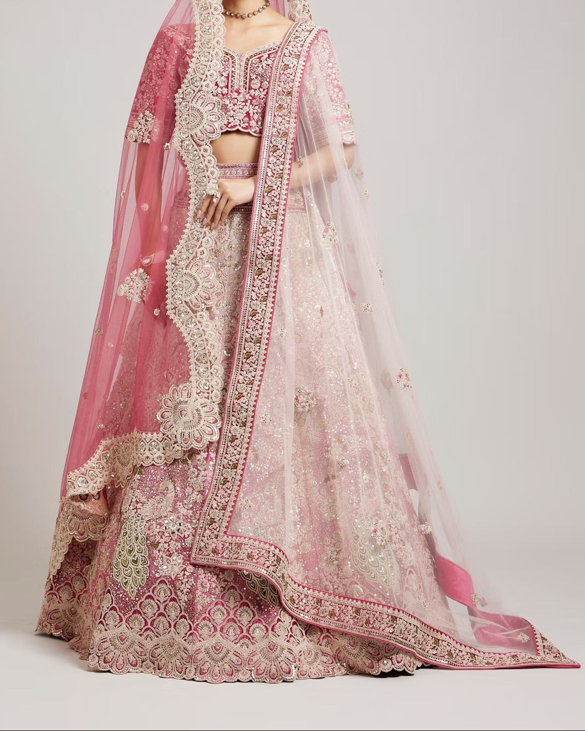 A woman modeling a heavily embroidered pink and blush bridal lehenga choli set with a sheer dupatta, photographed against a neutral light gray studio background.