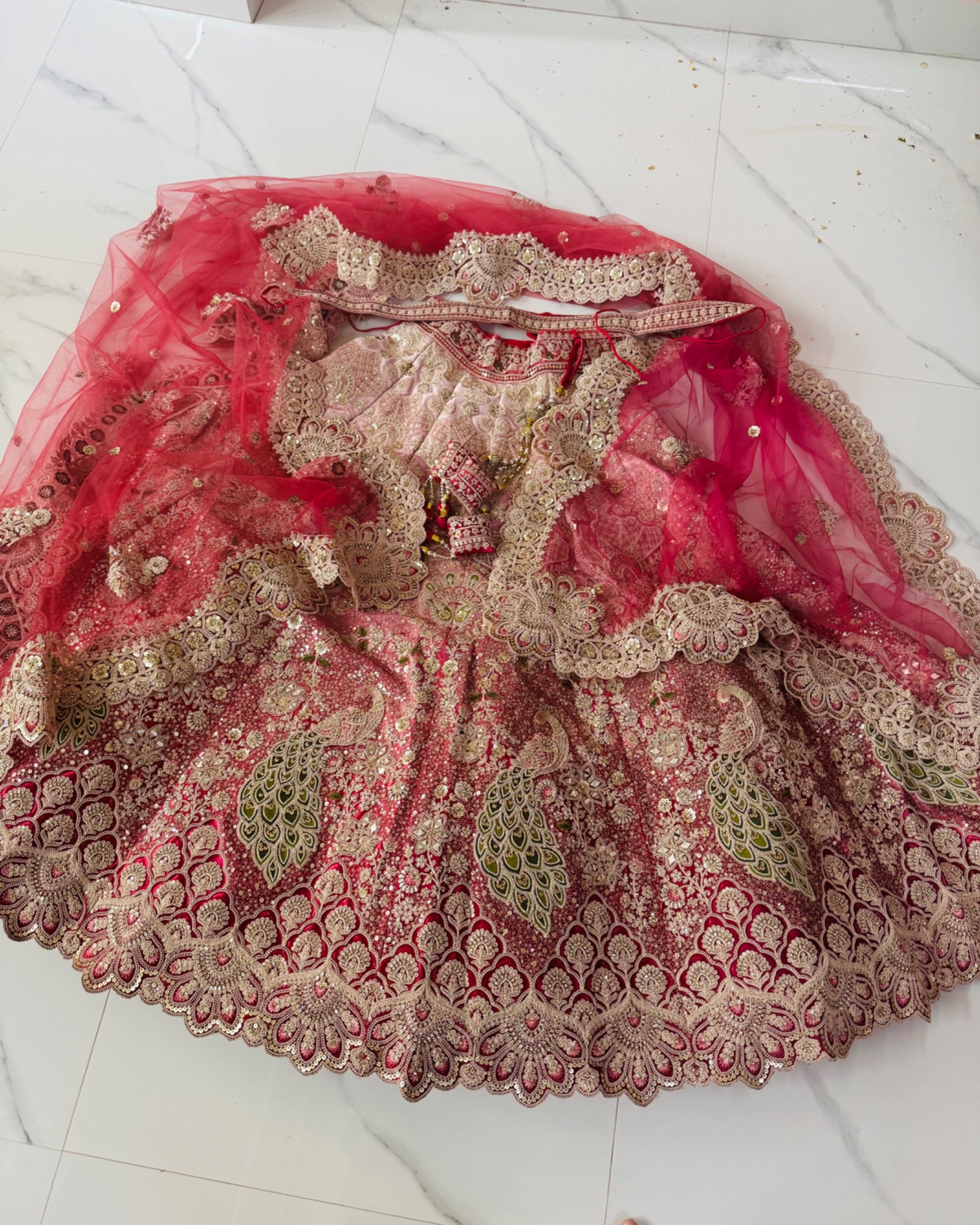 An ornate pink and gold embroidered bridal lehenga with a matching sheer pink dupatta, laid flat on a white marble tile floor.