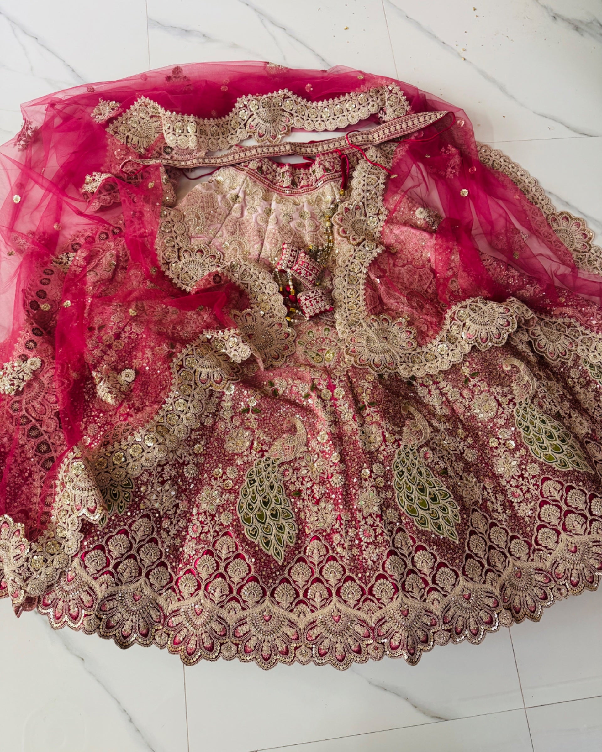 A heavily embroidered pink and beige traditional lehenga with a matching sheer pink dupatta, laid flat on a light marble surface. The fabric features intricate gold zari embroidery, sequins, and mirror work.