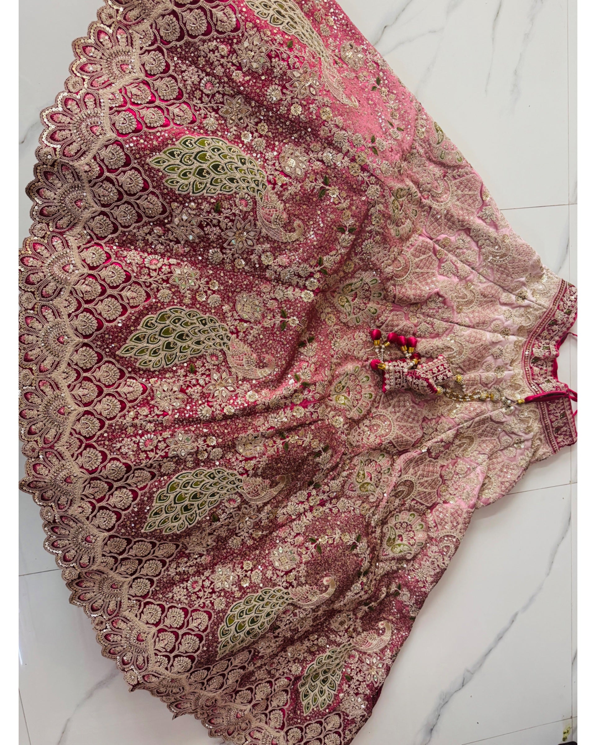 A pink and beige embroidered traditional skirt with intricate metallic gold and green mirror work, laid flat on a white marble floor.