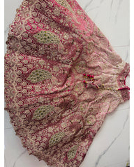 A pink and beige embroidered traditional skirt with intricate metallic gold and green mirror work, laid flat on a white marble floor.