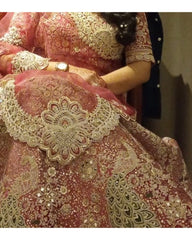 A close-up of a heavily embroidered traditional outfit in dusty pink and gold, featuring intricate lace, mirror work, and floral detailing. A person is partially visible wearing the garment, with one hand holding up a matching lace sleeve.