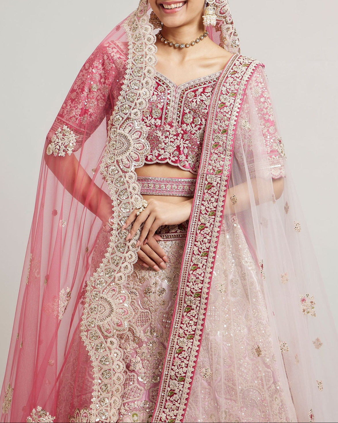 A woman modeling a traditional Indian bridal lehenga choli set in pink and light pink, heavily embroidered with gold and silver threadwork, paired with a sheer pink dupatta and jewelry against a plain light gray background.
