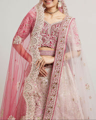 A woman modeling a traditional Indian bridal lehenga choli set in pink and light pink, heavily embroidered with gold and silver threadwork, paired with a sheer pink dupatta and jewelry against a plain light gray background.