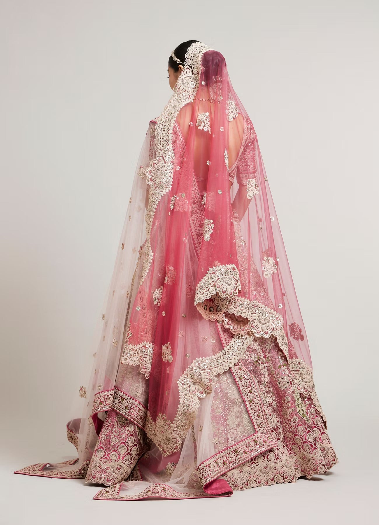 A woman shown from the back wearing an ornate pink and beige embroidered traditional bridal outfit with a sheer veil, photographed against a plain light gray studio background.