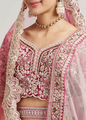A close-up of a pink embroidered traditional bridal outfit with a heavily embellished floral blouse, matching skirt, sheer dupatta, and ornate jewelry against a light gray background.