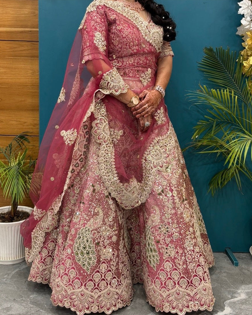A woman models an ornate pink and beige embroidered traditional outfit with a matching sheer dupatta, standing against a teal wall with potted plants and wood paneling.