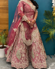 A woman models an ornate pink and beige embroidered traditional outfit with a matching sheer dupatta, standing against a teal wall with potted plants and wood paneling.