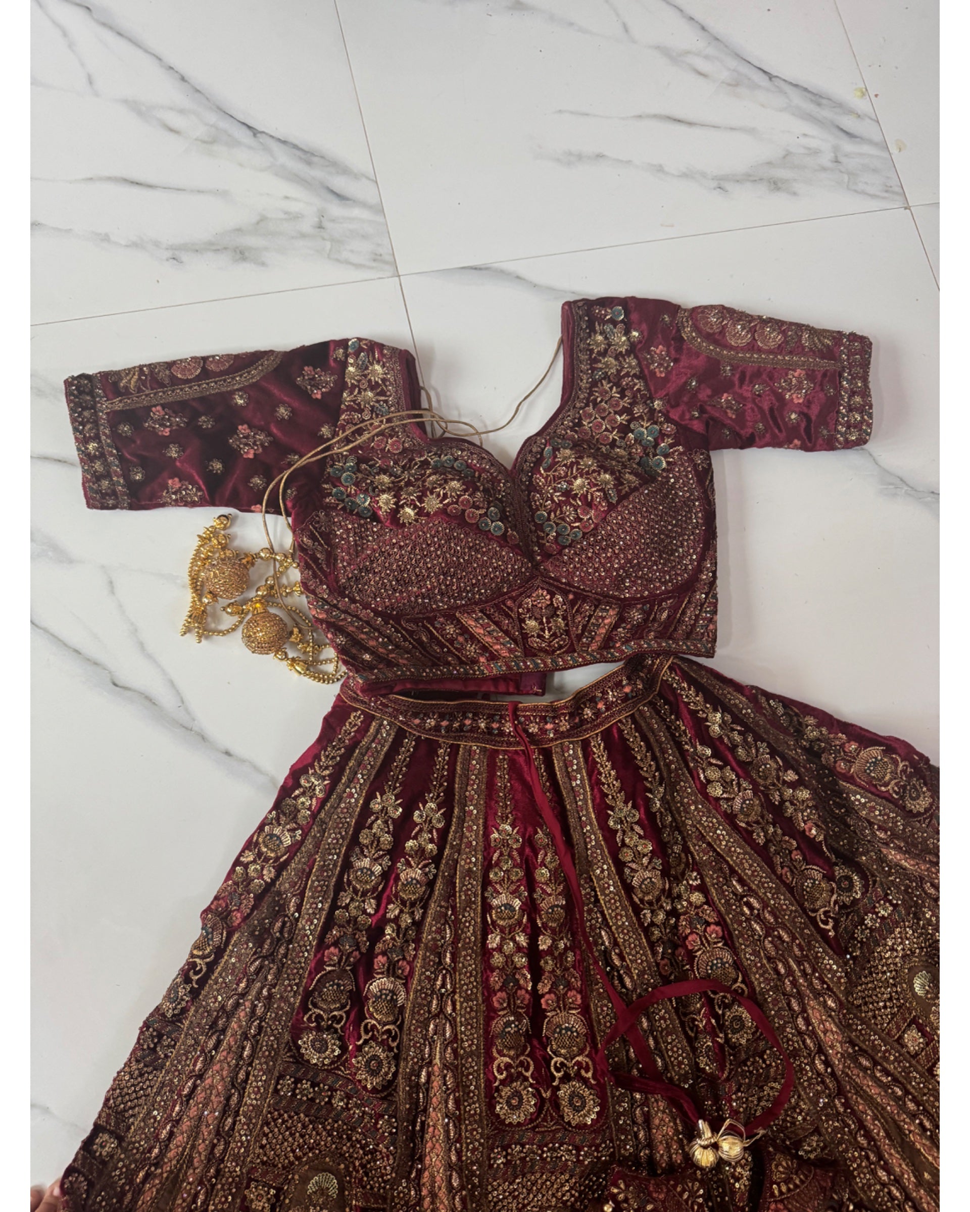 A traditional Indian lehenga choli set in deep maroon velvet with intricate gold and silver embroidery, laid flat on a white marble tiled floor. The set includes a heavily embellished sweetheart-style blouse, a matching pleated skirt with ornate border work, a gold-toned necklace, and a small matching clutch.