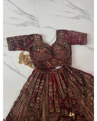 A traditional Indian lehenga choli set in deep maroon velvet with intricate gold and silver embroidery, laid flat on a white marble tiled floor. The set includes a heavily embellished sweetheart-style blouse, a matching pleated skirt with ornate border work, a gold-toned necklace, and a small matching clutch.