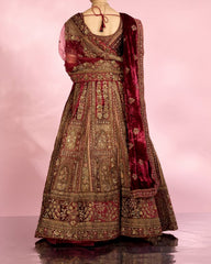 A woman stands with her back to the camera wearing an ornate traditional Indian lehenga choli set. The outfit is deep maroon red with extensive gold embroidery and features a matching sheer red blouse and dupatta, set against a soft pink studio backdrop.