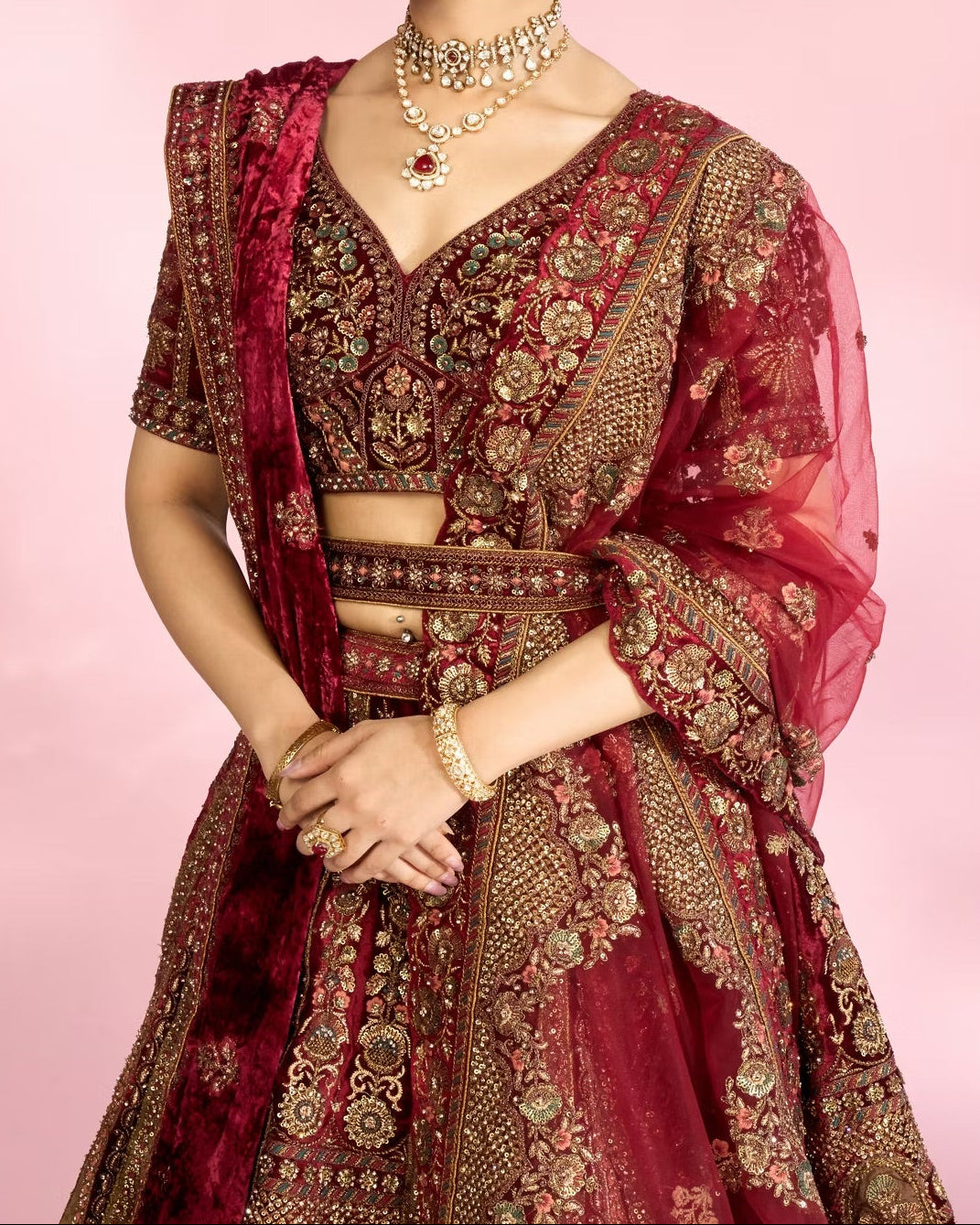 Woman in a traditional maroon and gold embroidered outfit with a pink background