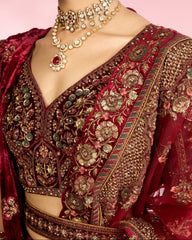 Close-up of a model wearing an ornate deep red and gold embroidered traditional blouse with intricate metallic floral patterns, paired with a matching sheer red dupatta and layered gold necklaces against a soft pink background.