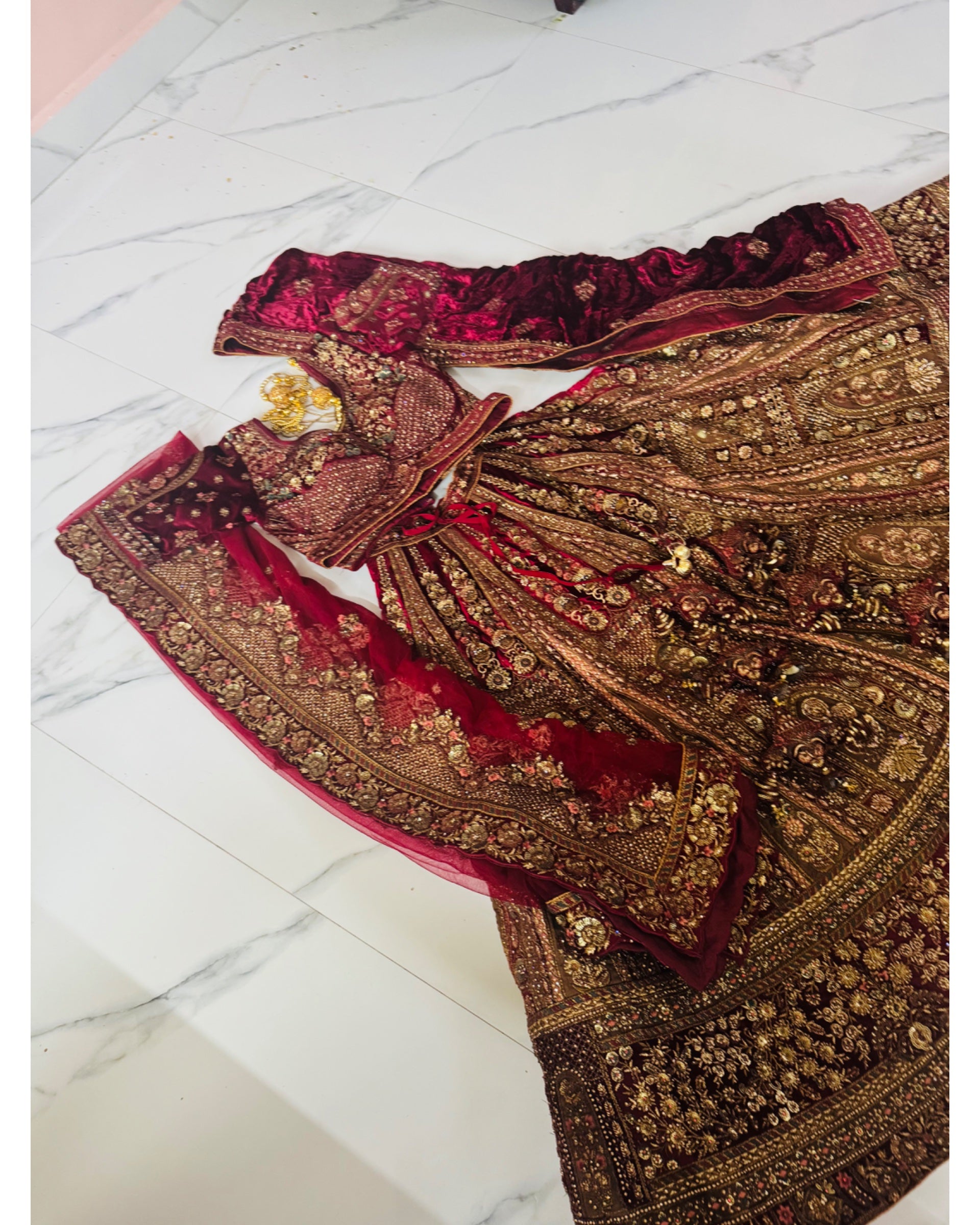 An ornate traditional bridal outfit laid flat on a white marble floor, featuring a heavily embroidered brown and gold lehenga skirt, a deep red velvet blouse with gold embroidery, matching red dupatta, and a red embroidered border piece.