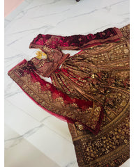 An ornate traditional bridal outfit laid flat on a white marble floor, featuring a heavily embroidered brown and gold lehenga skirt, a deep red velvet blouse with gold embroidery, matching red dupatta, and a red embroidered border piece.