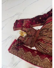 A deep red velvet and nette traditional Indian outfit with intricate gold embroidery, laid out on a white marble floor. The set includes a heavily embellished kurta, matching skirt, and a coordinating red dupatta.