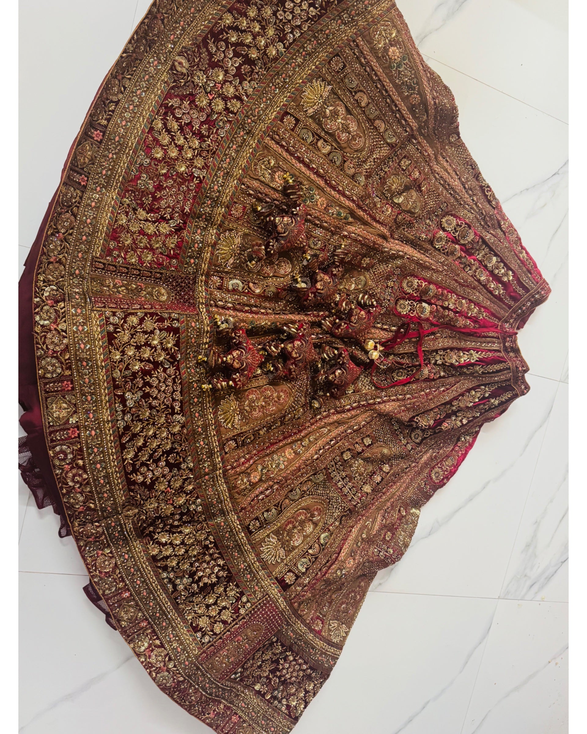 An ornate traditional skirt or lehenga laid flat on a light marble floor, featuring intricate gold zari embroidery, red and gold floral patterns, and heavy bead embellishments.
