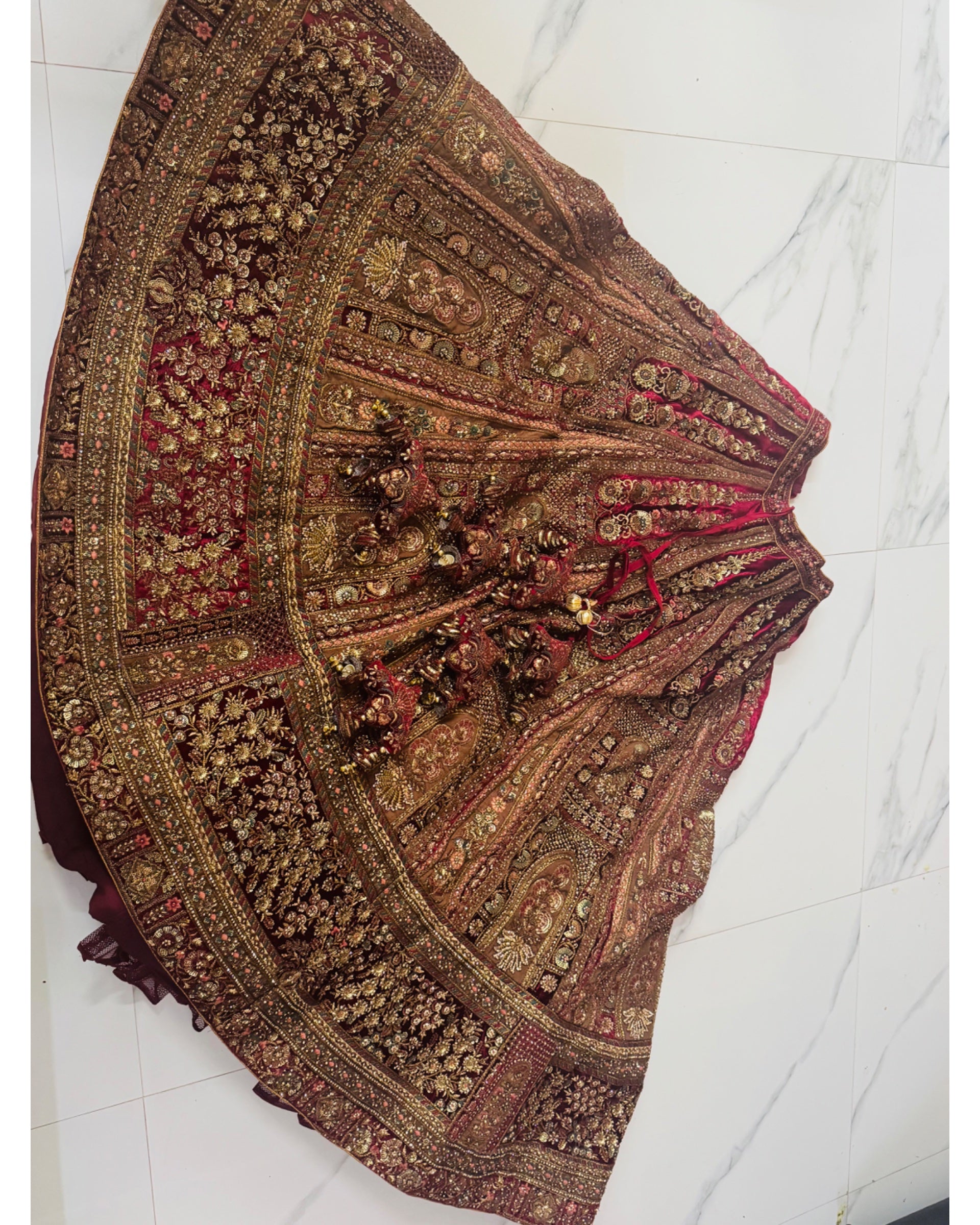 An ornate, heavily embroidered traditional skirt or lehenga laid flat on a white marble tile floor. The fabric is primarily brown with intricate gold zari embroidery, red inner lining, and detailed beadwork.
