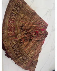 An ornate, heavily embroidered traditional skirt or lehenga laid flat on a white marble tile floor. The fabric is primarily brown with intricate gold zari embroidery, red inner lining, and detailed beadwork.