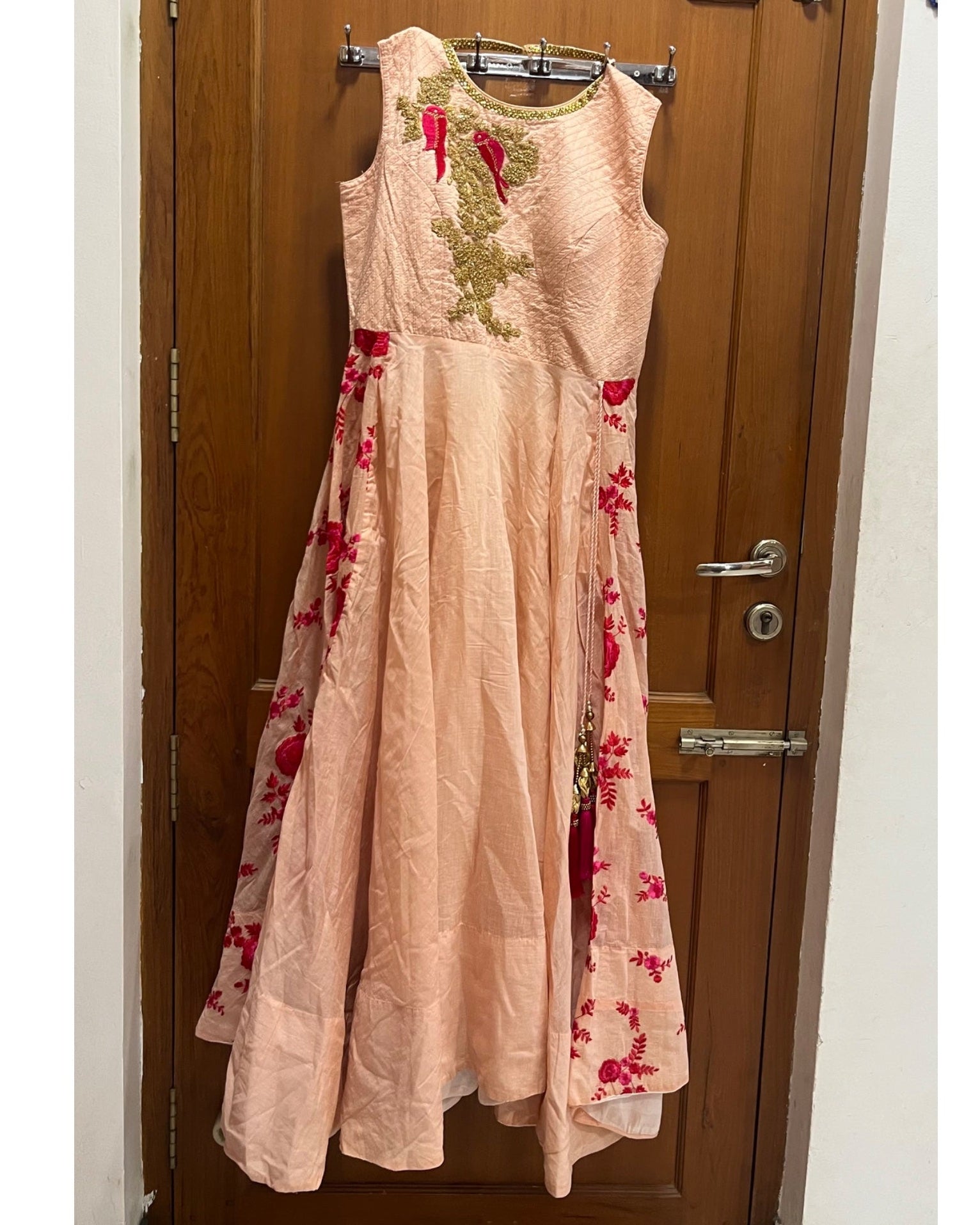 A sleeveless peach-pink maxi dress with red floral embroidery and gold mirror-work embroidery on the bodice, hanging on a metal hanger in front of a brown wooden door.