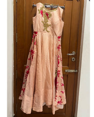 A sleeveless peach-pink maxi dress with red floral embroidery and gold mirror-work embroidery on the bodice, hanging on a metal hanger in front of a brown wooden door.