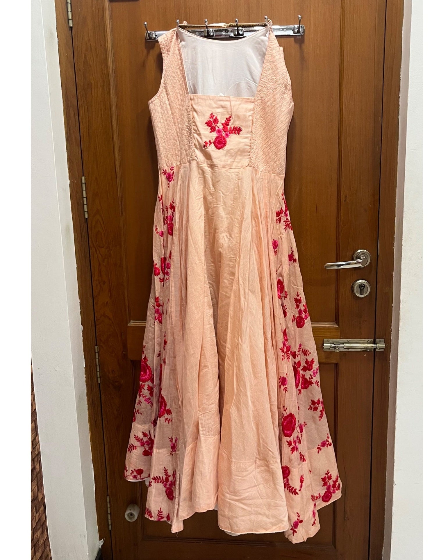 Back view of a sleeveless peach maxi dress with red floral embroidery and a white inner lining, hanging on a clothes rack in front of a wooden door.