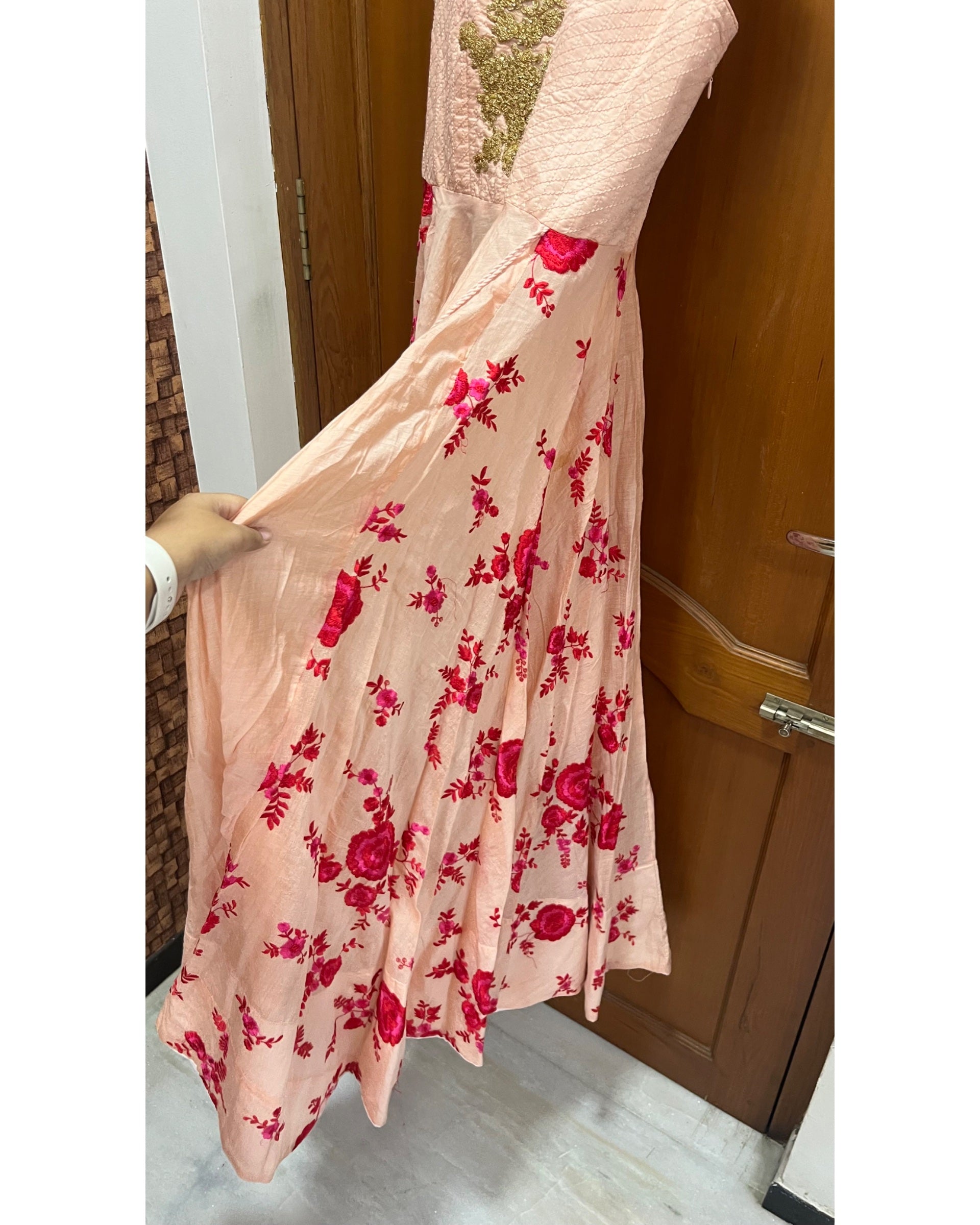 A peach-colored maxi dress with a vibrant red floral print and a gold embroidered bodice, held up by a hand in an indoor setting.