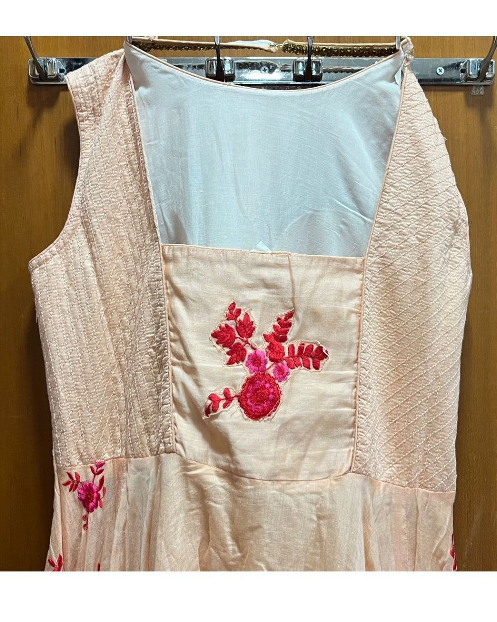 A sleeveless light beige dress with pink floral embroidery on the front pocket, hanging on a metal hanger against a wooden door.