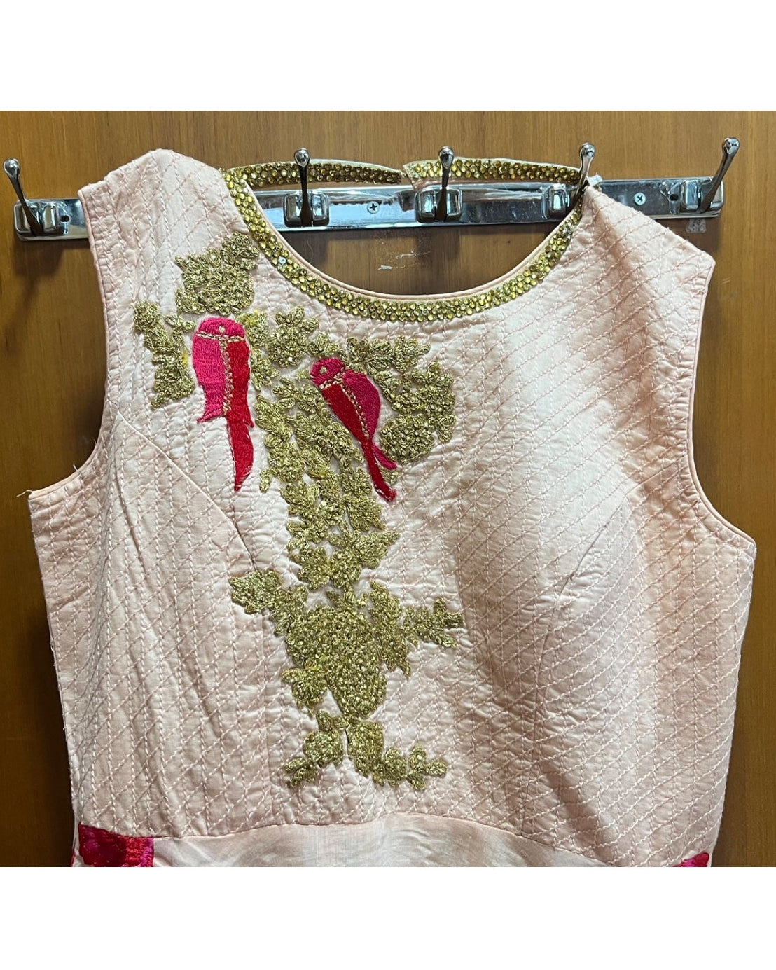 A sleeveless light pink or beige embroidered top hanging on a metal rack against a wooden door. The top features olive green floral embroidery and two bright pink embroidered motifs on the chest.