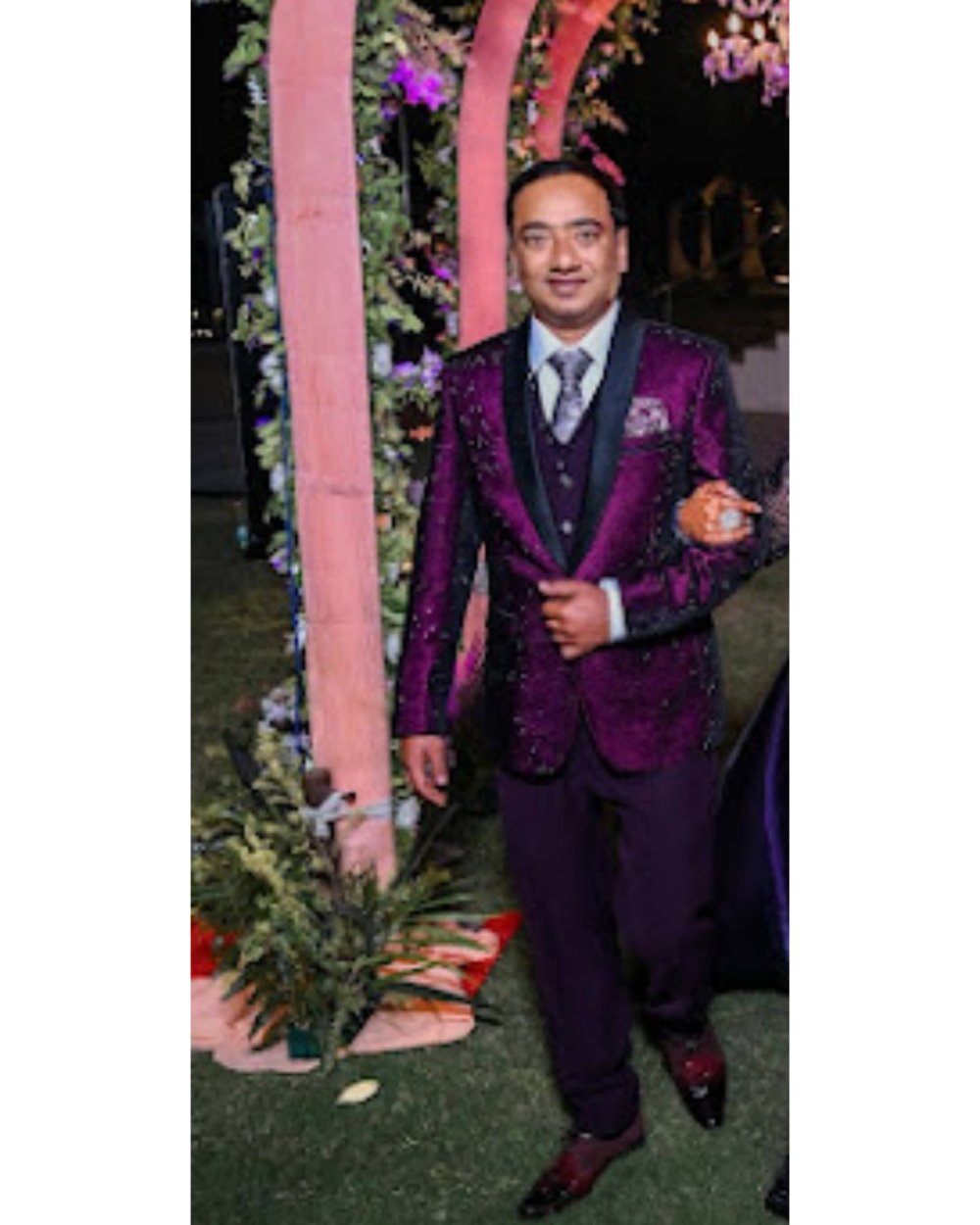 A man wearing a sparkling purple sequined blazer with matching purple trousers, a black vest, and a patterned tie, standing in front of a pink floral arch at night.