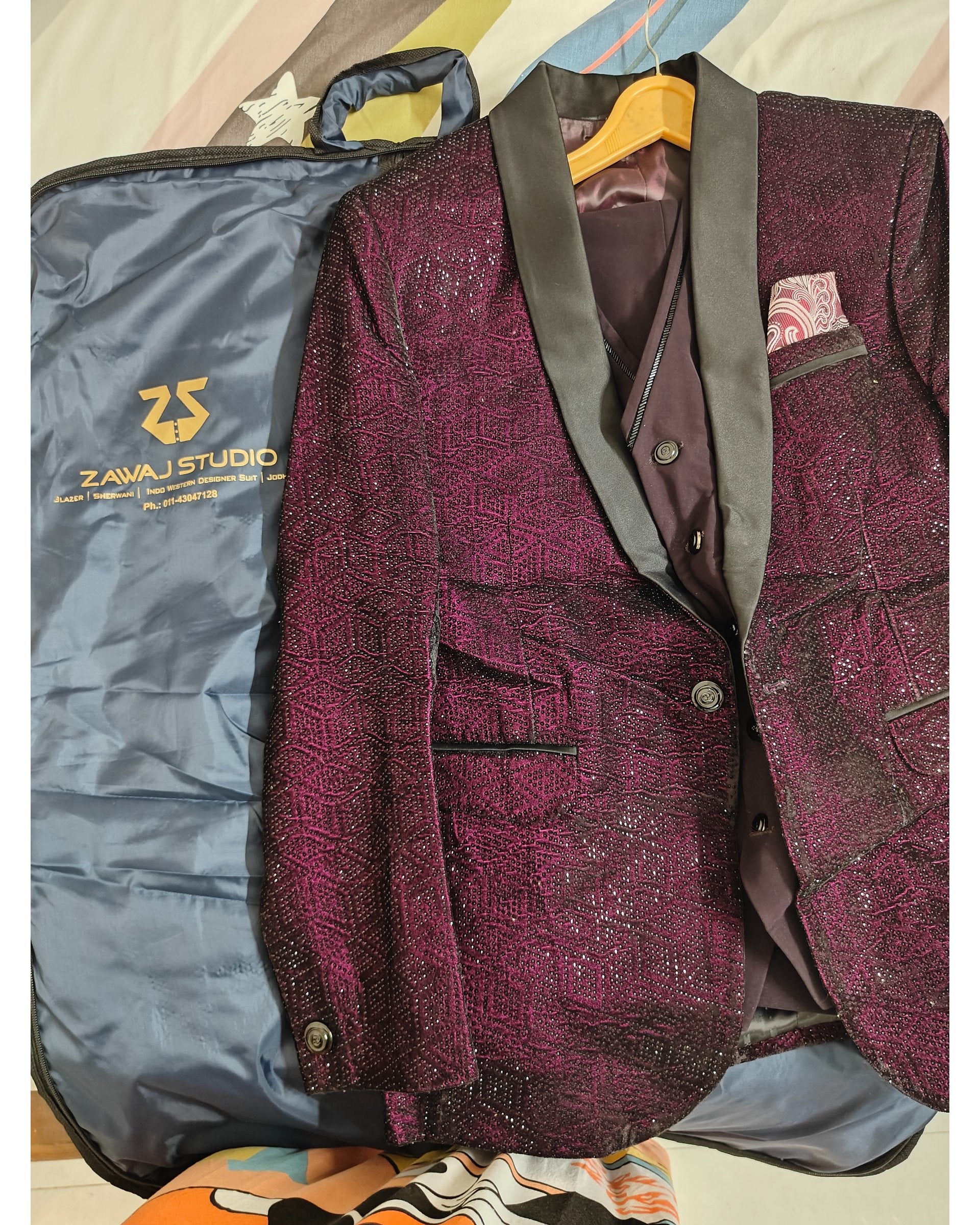 A men's tailored blazer in deep burgundy with a textured pattern, dark gray satin shawl collar, and a matching dark waistcoat, laid flat next to its blue garment bag. The brand 'Zawaj Studio' is visible on the garment bag.