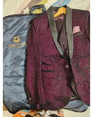 A men's tailored blazer in deep burgundy with a textured pattern, dark gray satin shawl collar, and a matching dark waistcoat, laid flat next to its blue garment bag. The brand 'Zawaj Studio' is visible on the garment bag.