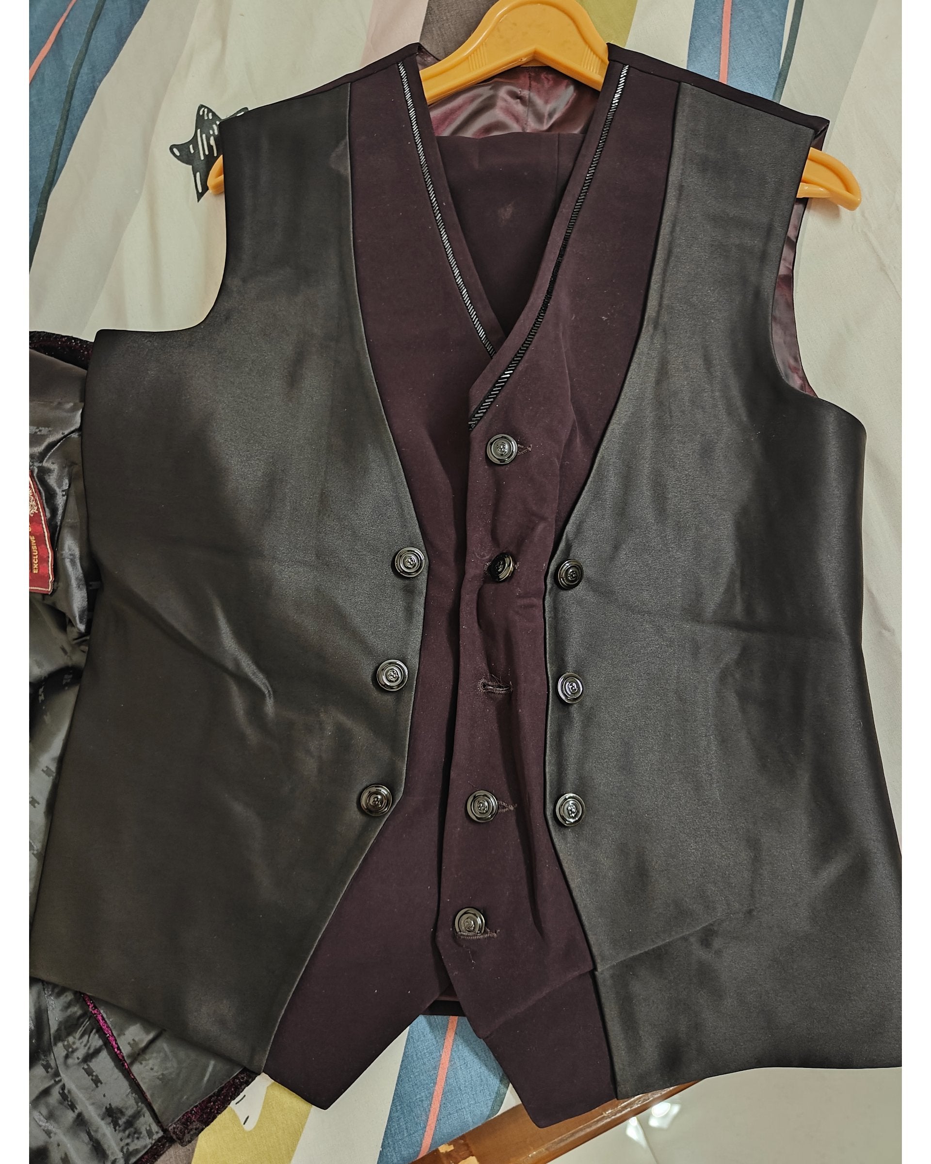 A men's double-breasted waistcoat vest in black satin, layered over a matching dark plum purple vest, hanging on a wooden hanger.