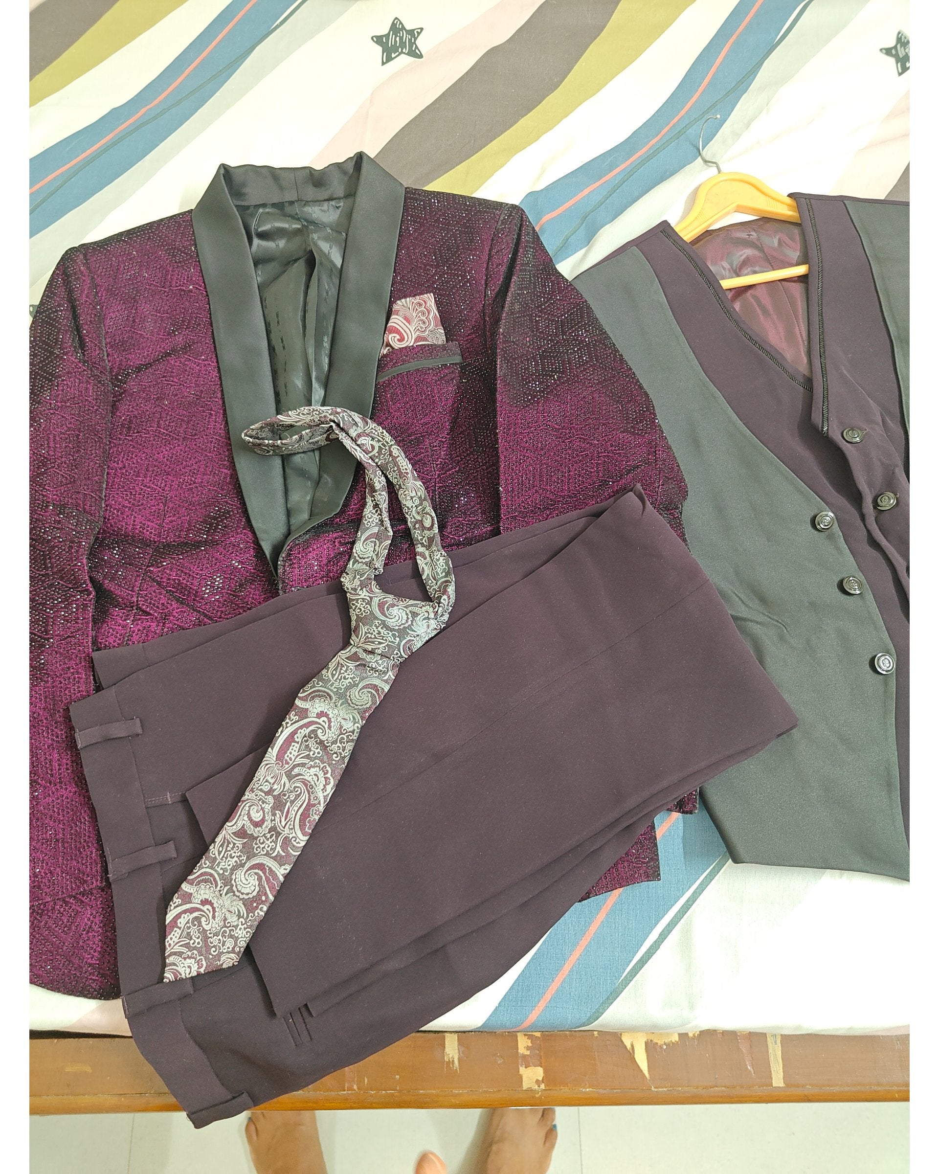 A men's formal suit set laid out on a striped sheet, including a textured purple blazer with dark green satin lapels, matching dark purple trousers, a dark gray vest on a wooden hanger, and a paisley patterned tie.