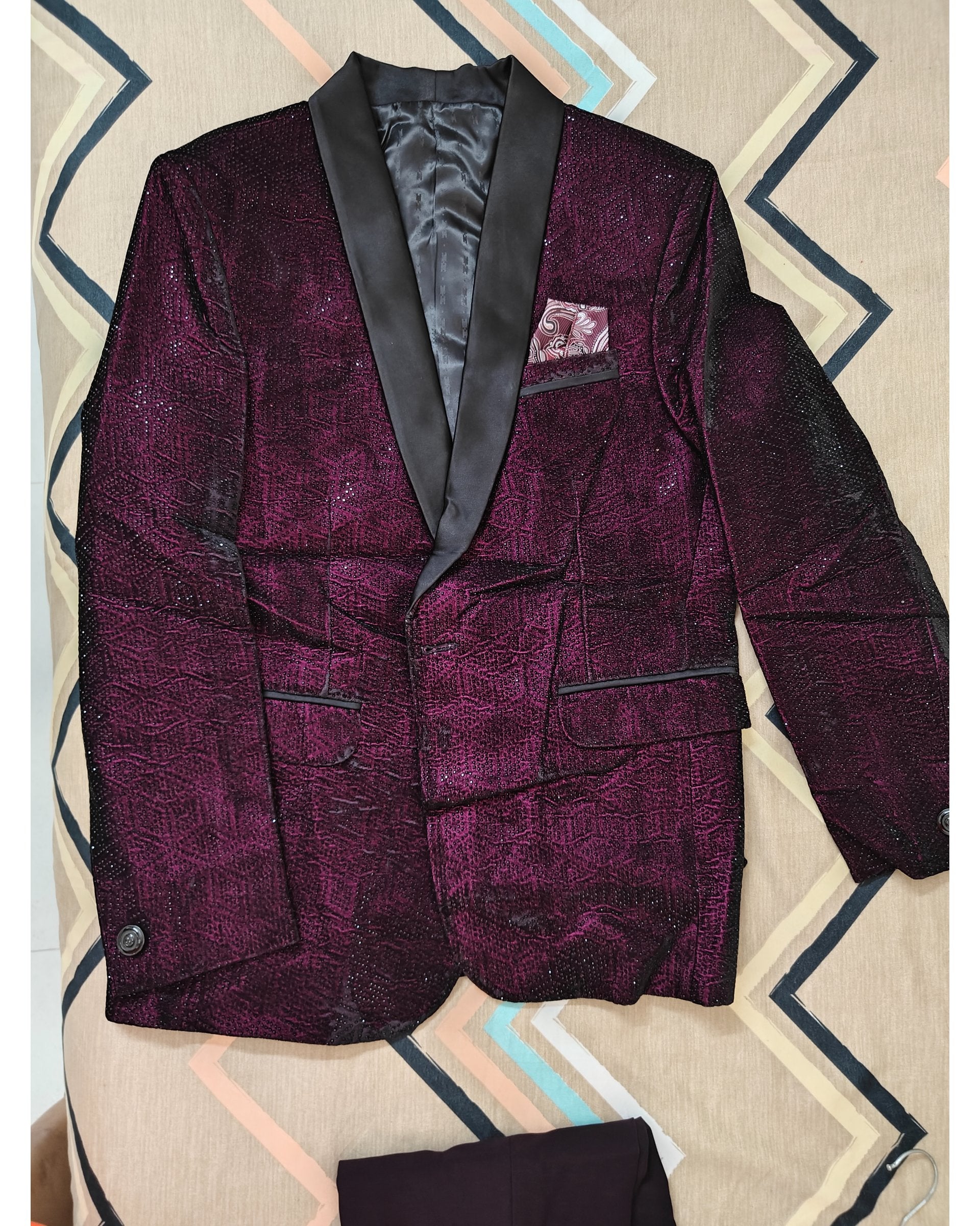A men's formal blazer in a deep purple brocade pattern with black satin shawl lapels, displayed flat on a patterned beige and blue background.