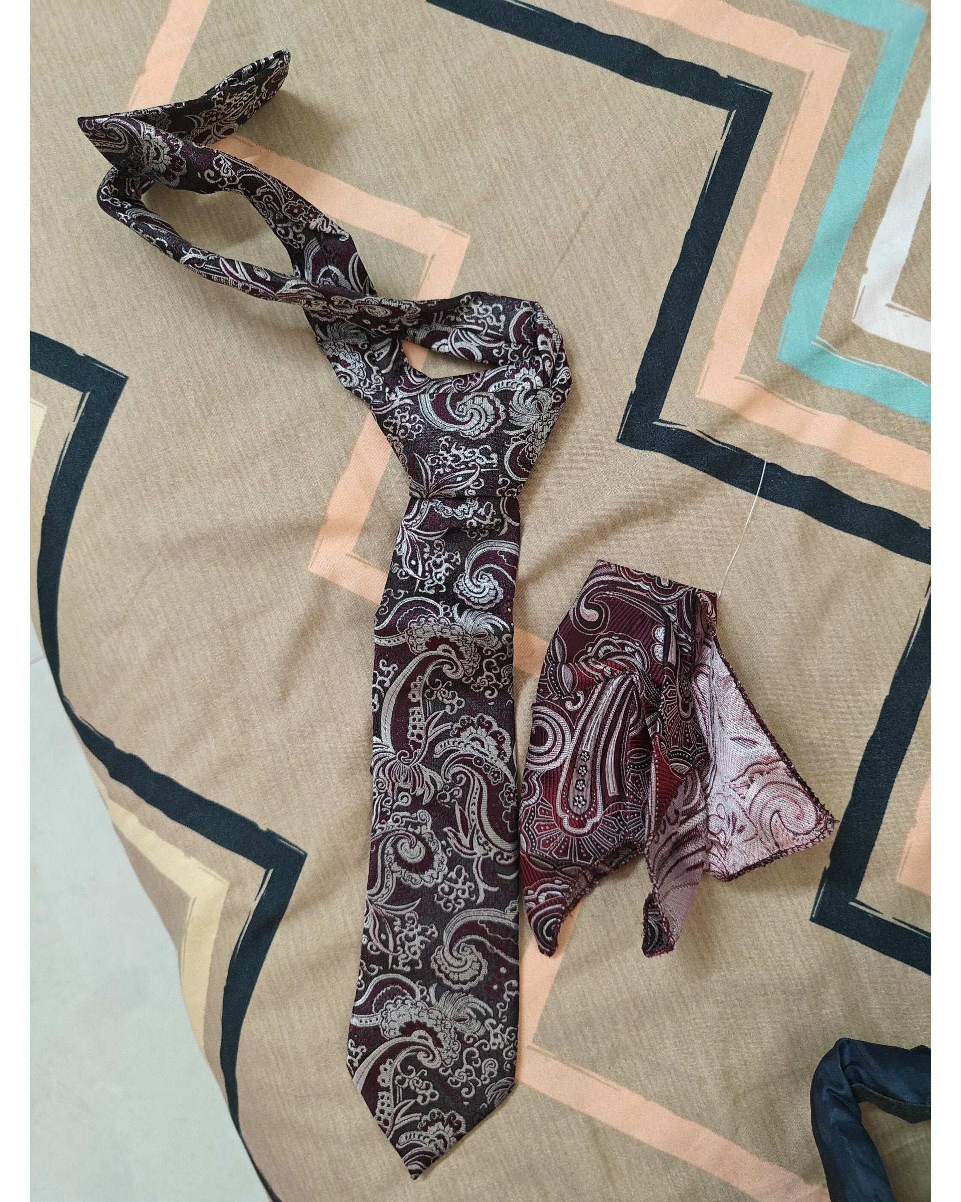 A men's necktie and matching pocket square set with a dark brown, burgundy, and silver paisley pattern, laid out on a beige and blue geometric patterned fabric background.