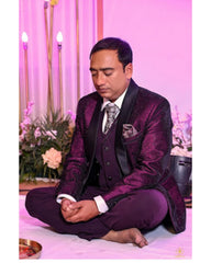 A man wearing a dark purple and black three-piece suit with a textured patterned jacket, sitting cross-legged on a white floor against a pink backdrop with decorative flowers and candles.