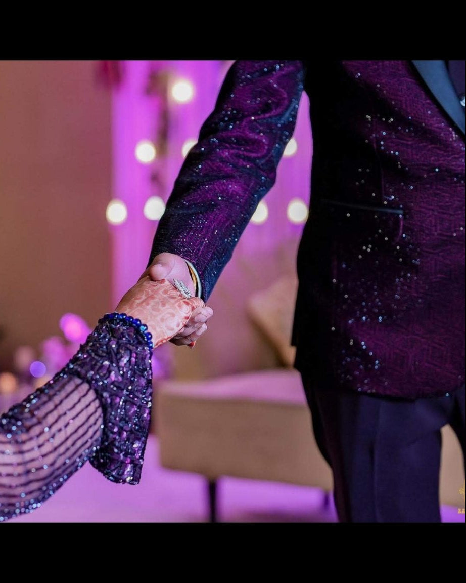 A close-up of a person in a sparkling dark purple sequined jacket holding hands with another person wearing a sheer purple sleeve, set against a softly blurred background with pink and white lights.