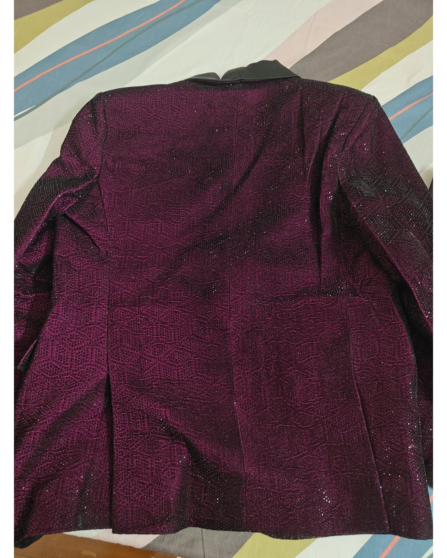 A dark plum purple textured fabric garment, back view of jacket, laid flat on a striped background.
